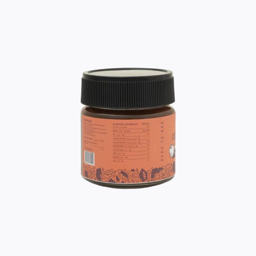A small amber glass jar with a black ribbed screw cap and an orange label, shown from the back on a plain white background.