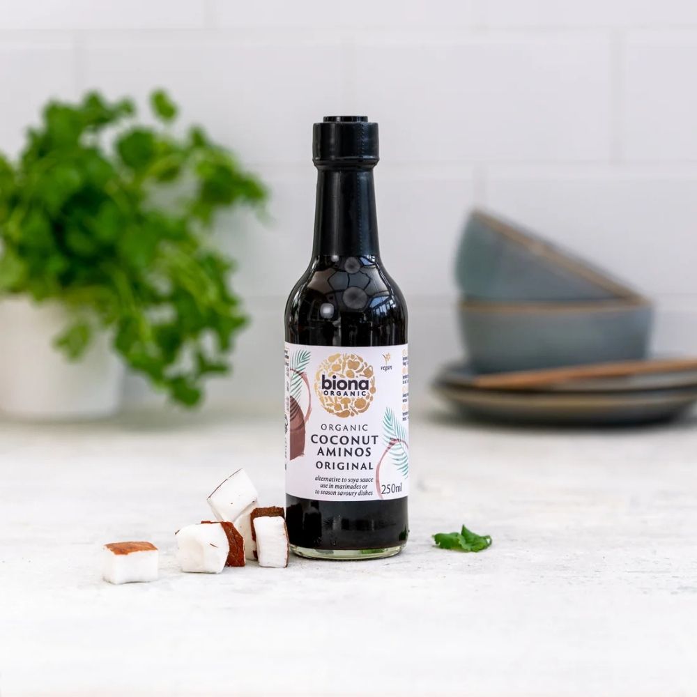 Biona Organic Coconut Aminos bottle on a white surface with a plant and bowls in the background