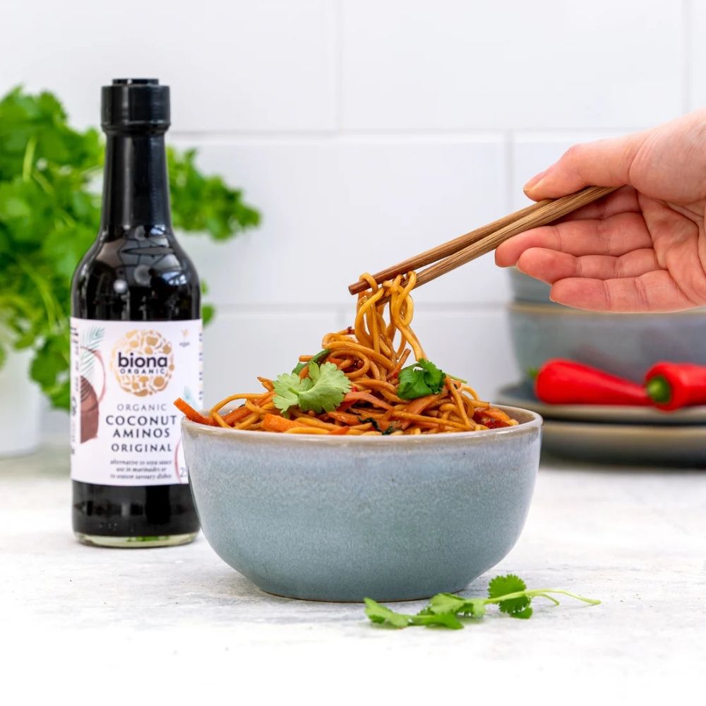 Bowl of noodles with chopsticks, bottle of Biona coconut aminos, and red peppers on a white background