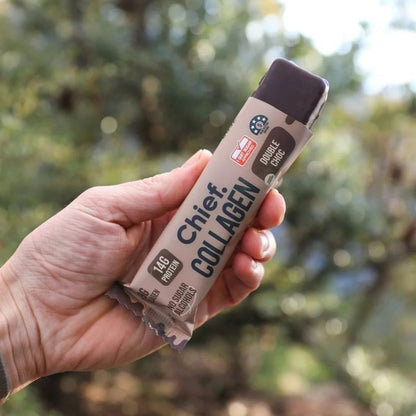 A hand holding a wrapped collagen bar outdoors, with a blurred natural background of green foliage and bright light. The wrapper is beige with dark brown and black text reading 'Chief Collagen' and 'Double Choc'.