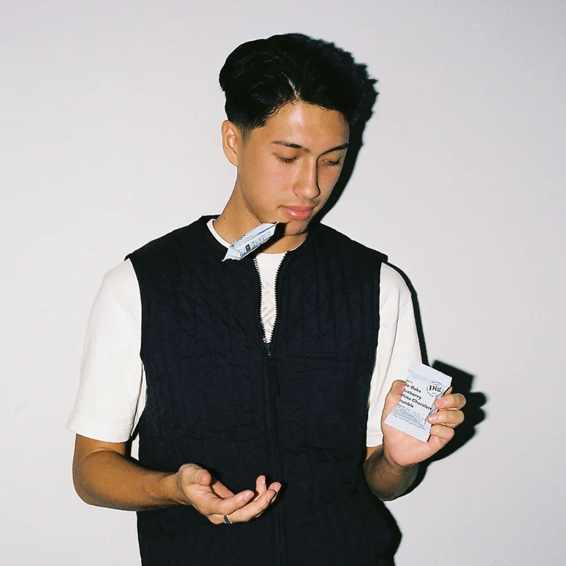 A young man with dark hair looking down while holding a small white packet in his right hand and gesturing with his left hand. He is wearing a black quilted zip-up vest over a white t-shirt, standing against a plain off-white wall.