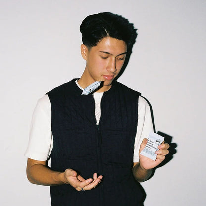 A young man with dark hair looking down while holding a small white packet in his right hand and gesturing with his left hand. He is wearing a black quilted zip-up vest over a white t-shirt, standing against a plain off-white wall.