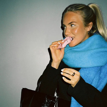 A woman with blonde hair in a ponytail wearing a bright blue knit scarf and a black coat, eating a pink pastry. She is holding a black glossy bag against a plain gray background.