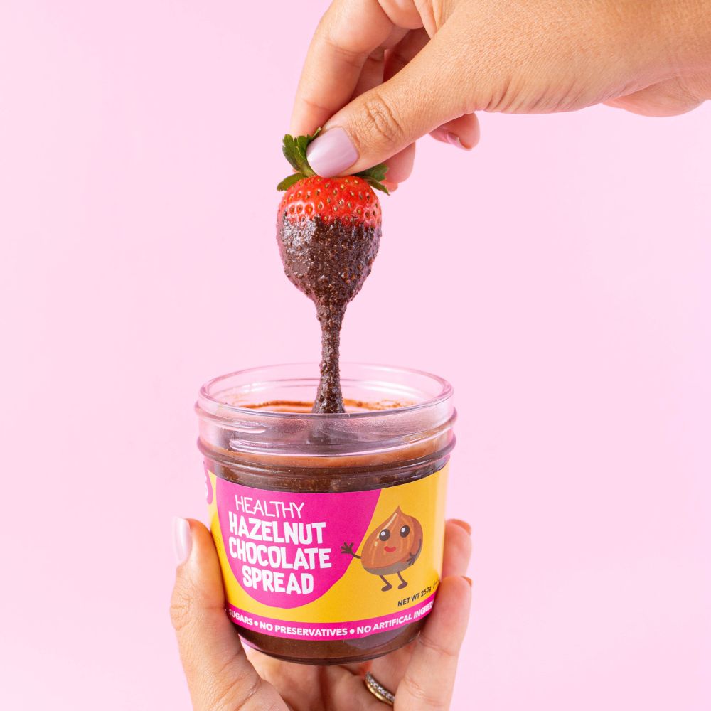Hand dipping a strawberry into a jar of healthy hazelnut chocolate spread against a pink background