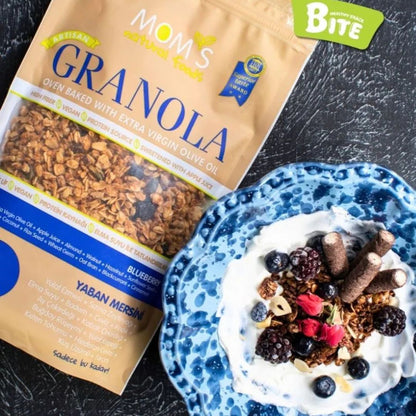 Mom's Granola package next to a bowl of granola with yogurt and berries on a dark surface