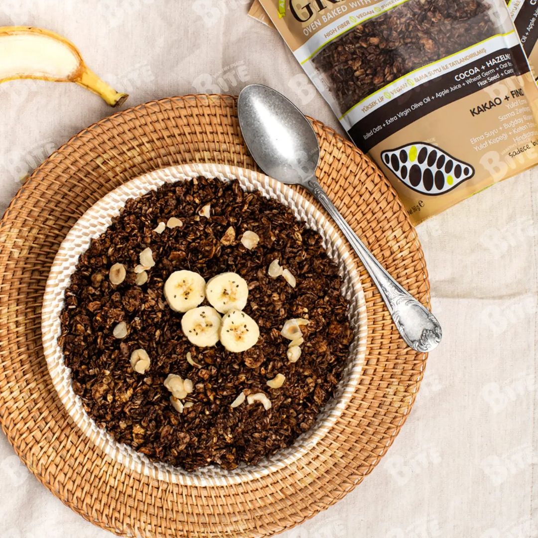 Bowl of granola with bananas and a spoon on a woven mat, with a package of Granola in the background.