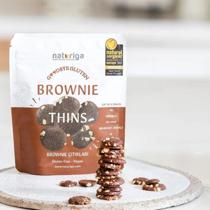 A stand-up pouch of Naturiga Goodbye Gluten Brownie Thins sits on a light stone tray with several brownie cookies placed in front. The package is white and brown with a Natural Organic Awards 2021 badge, and the cookies are shown as round chocolate-dusted pieces with visible nut pieces.