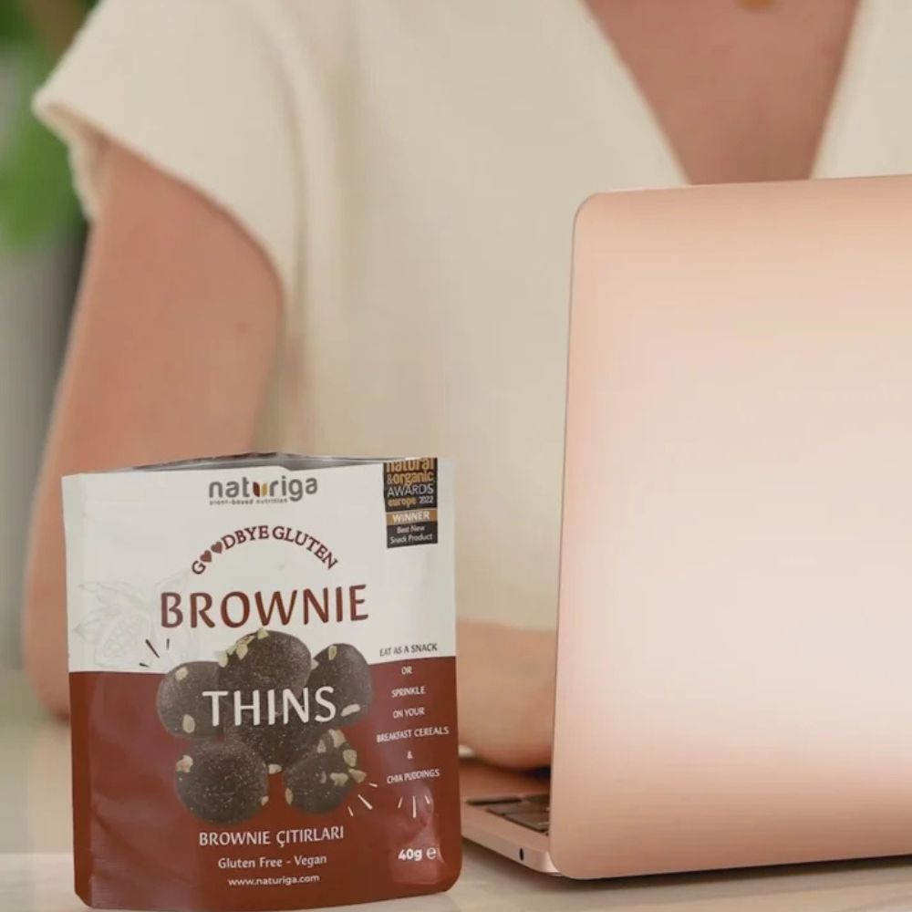 A box of Naturiga Goodbye Gluten Thins Brownie (Cıtbları) gluten free protein powder snacks is shown on a white surface next to an open rose gold laptop, with a person in a white top blurred in the background.