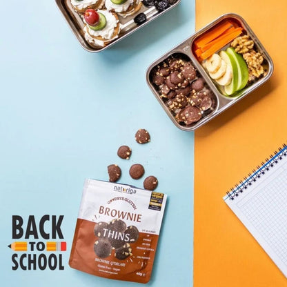A styled flat-lay showing a bag of Nateriga Brownie Thins on a split light blue and orange background, with loose chocolate pieces scattered nearby and two metal lunch boxes containing snacks. A 'BACK TO SCHOOL' graphic appears in the lower left.