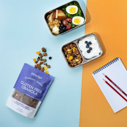 A bag of Nalwriqa gluten free granola is displayed on a split light blue and orange background, styled with a lunch box, a granola and yogurt bowl, and a notebook with pencils.