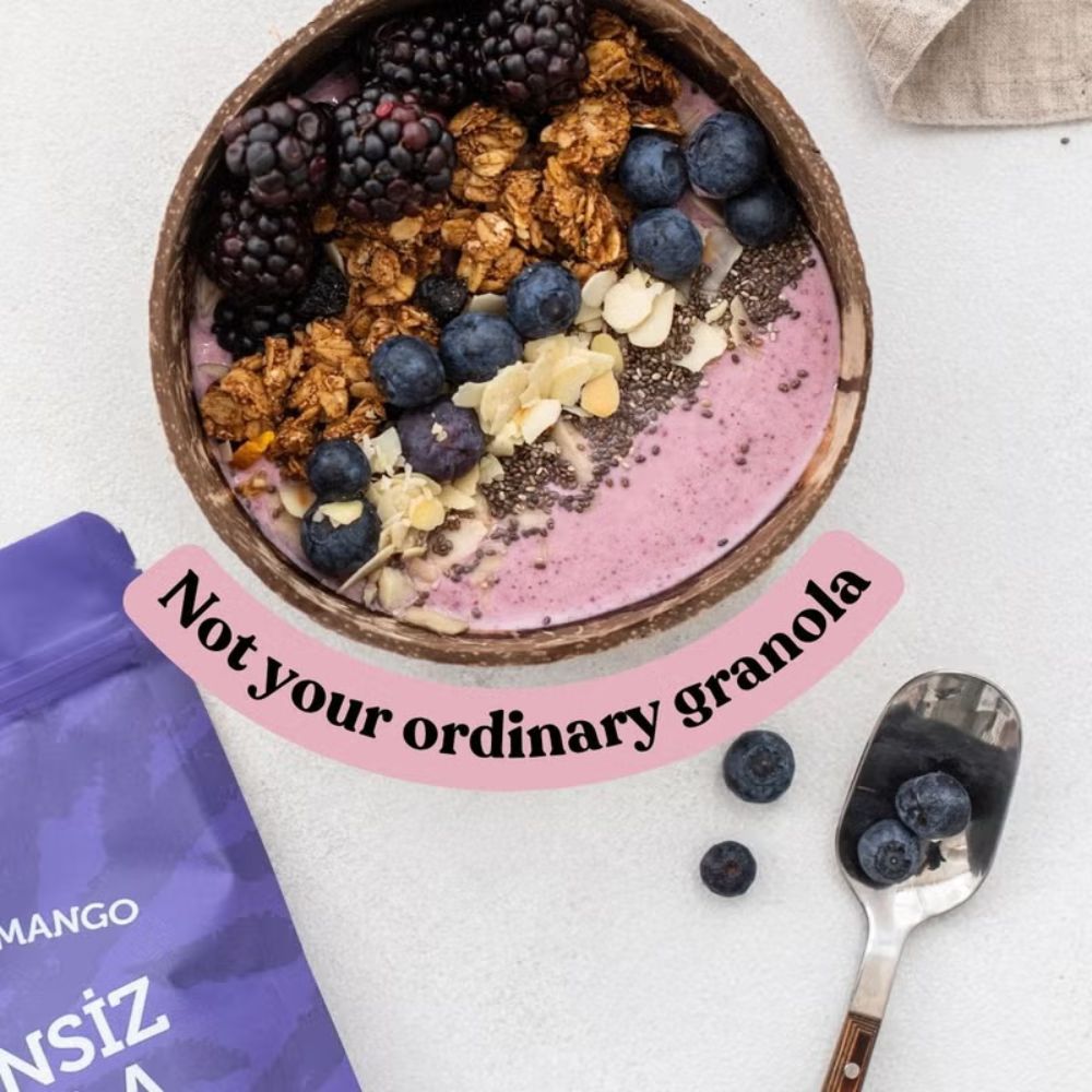 A bowl of pink smoothie topped with blackberries, blueberries, granola, and coconut flakes, styled on a light surface with a purple pouch, a spoon, and a text overlay reading 'Not your ordinary granola'.