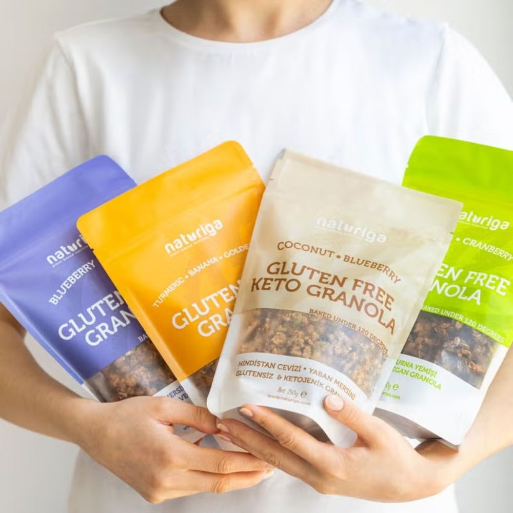 A person in a white shirt holds four Naturiga gluten-free keto granola pouches in different colors: blue, orange, beige, and green, against a clean white background.