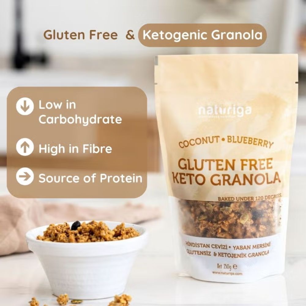 A stand-up pouch of naturiga Gluten Free Keto Granola in Coconut & Blueberry flavor, shown next to a small white bowl filled with the granola. The background is a softly blurred kitchen setting.
