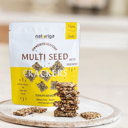 A package of Naturiga Multi Seed Crackers, Plain Sade flavor, displayed on a round stone plate with several crackers stacked and scattered in front. The background is a soft, blurred kitchen setting.
