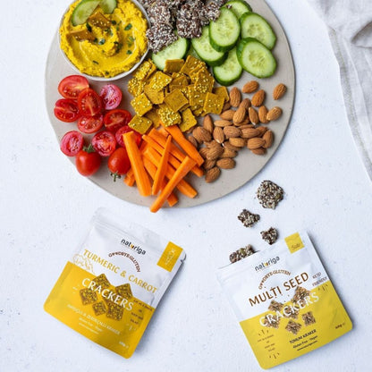 Two bags of netriuga crackers placed on a white surface below a styled snack board with hummus, cucumbers, tomatoes, carrots, almonds, and seeds.