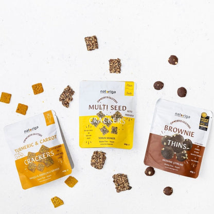 Three pouches of Naturliga organic crackers and multi-seed snacks are arranged on a white surface with scattered pieces of the crackers and chocolate chips around them. The pouches are labeled Turmeric & Carrot Crackers, Multi Seed Crackers, and Brownie Thins.