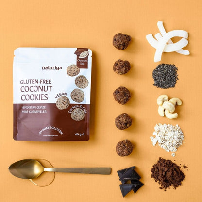 A flat-lay of a pouch of Naturiga Gluten-Free Coconut Cookies alongside the ingredients used to make them, arranged on a warm orange background. The ingredients include shredded coconut, black seeds, cashews, oats, cocoa powder, chocolate chunks, and a gold spoon.