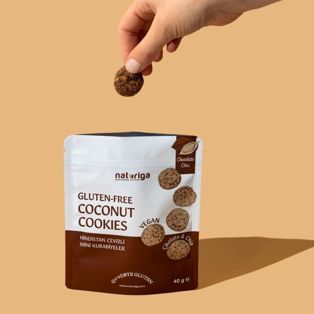 A hand holds a single round gluten-free coconut cookie above a stand-up pouch of Naturiga Gluten-Free Coconut Cookies on a warm beige background.