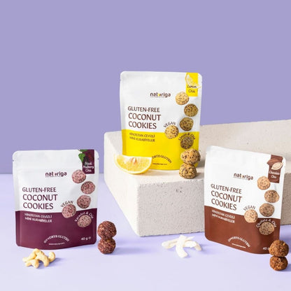 Three bags of naturiGA gluten-free coconut cookies arranged on a lavender studio background, with two bags standing upright on a white block and one bag placed in front. A lemon wedge and a few cookies are styled around the products.
