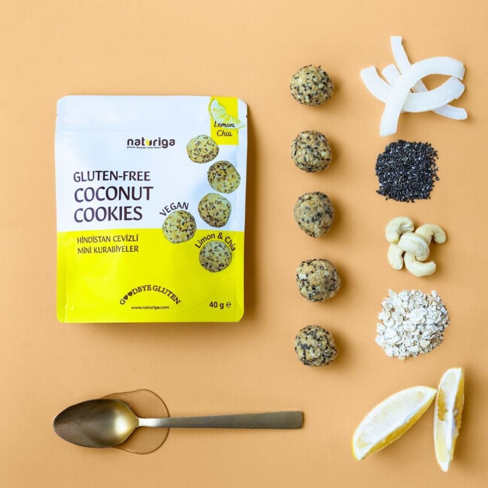 A flat-lay of a Natriva gluten-free coconut cookies pouch on a warm beige background, surrounded by ingredients used to make the cookies: lemon slices, cashews, chia seeds, coconut flakes, oats, and several mini cookies. A metal spoon is also shown.