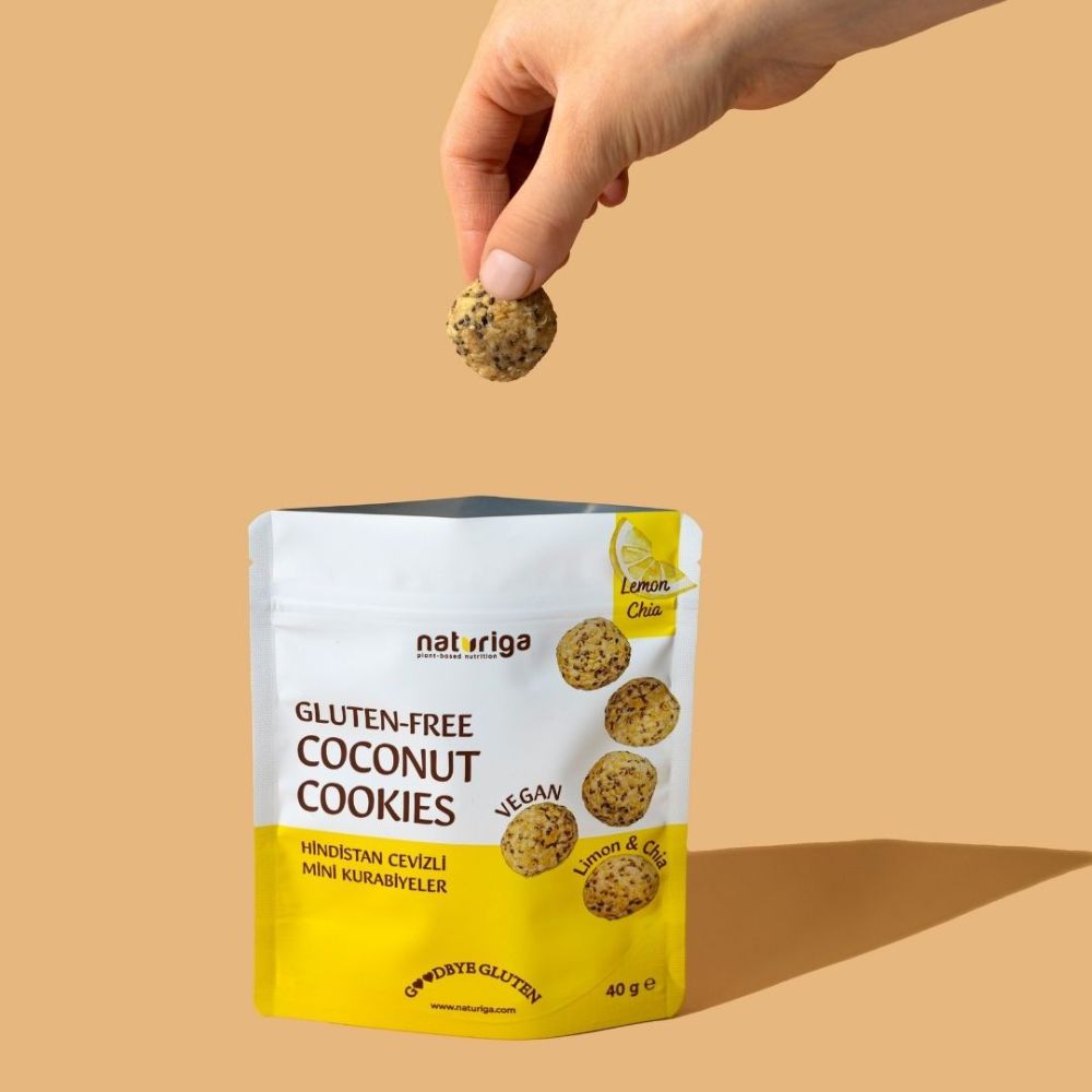 A hand holds a small round gluten-free coconut cookie above a white and yellow pouch of Naturiga Gluten-Free Coconut Cookies against a solid beige background.