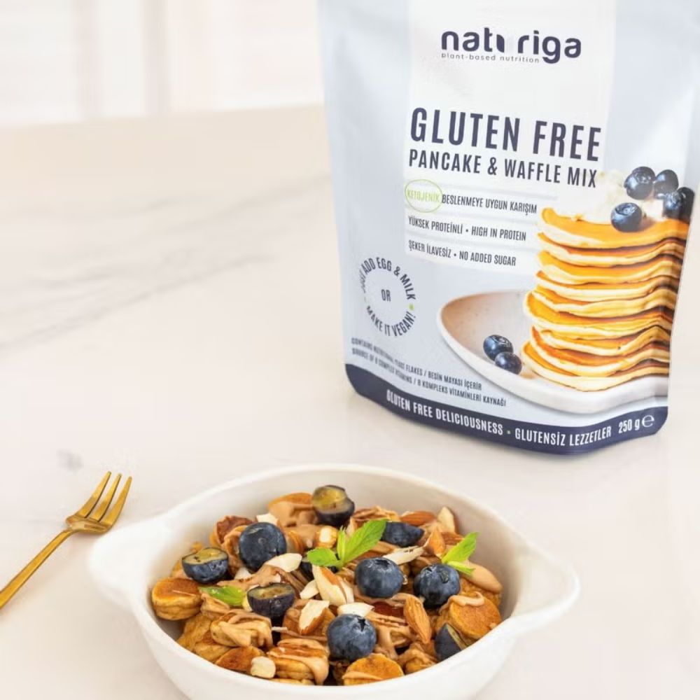 A bag of nat riga Gluten Free Pancake & Waffle Mix stands next to a white bowl filled with prepared pancakes topped with blueberries, almonds, and mint, on a light marble surface with a gold fork.