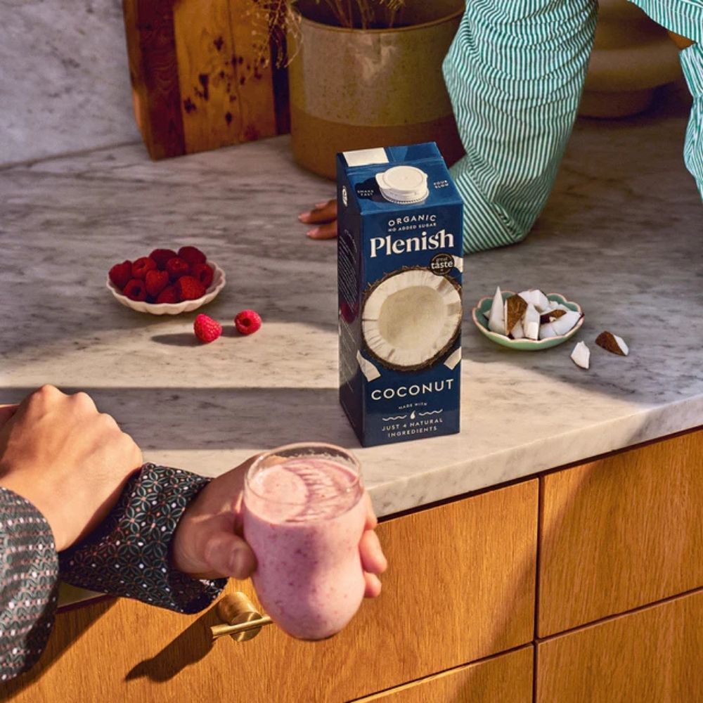 Person holding a glass of a pink smoothie next to a box of Plenish coconut milk on a kitchen counter.