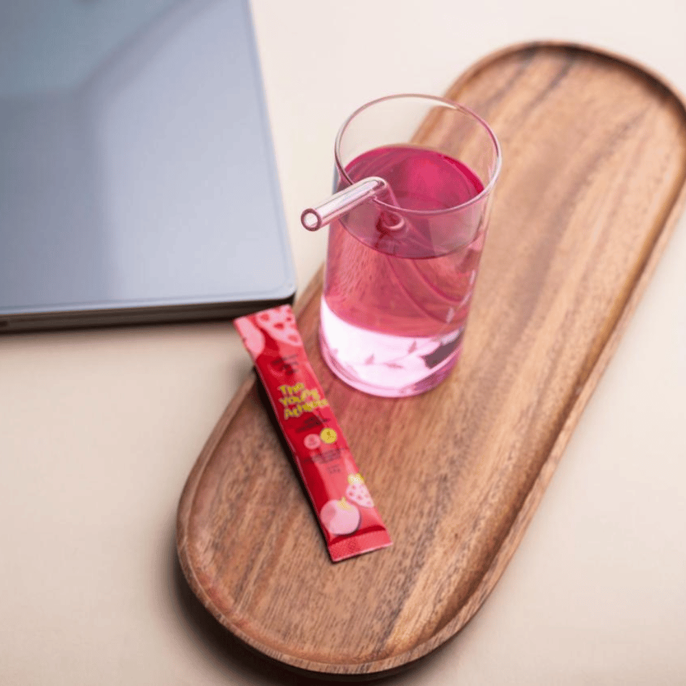 Pink drink in a glass with a straw on a wooden tray next to a red packet, with a laptop in the background.