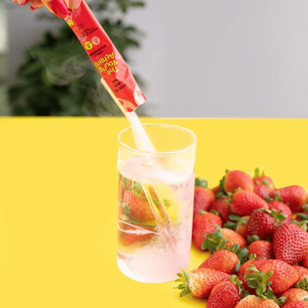 Strawberry-flavored drink mix being poured into a glass with strawberries on a yellow surface.