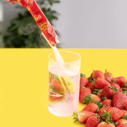Strawberry-flavored drink mix being poured into a glass with strawberries on a yellow surface.