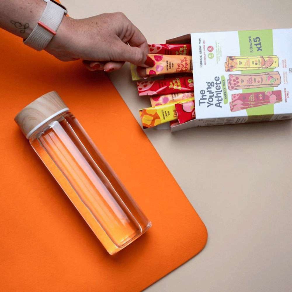 Clear glass bottle with wooden cap on an orange surface, next to a box of 'The Young Athlete' snack bars.