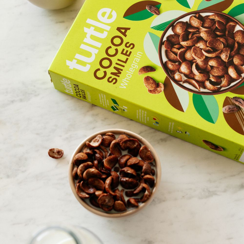 A box of Turtle Cocoa Smiles Wholegrain cereal is shown on a light marble surface next to a small bowl filled with the cereal pieces.