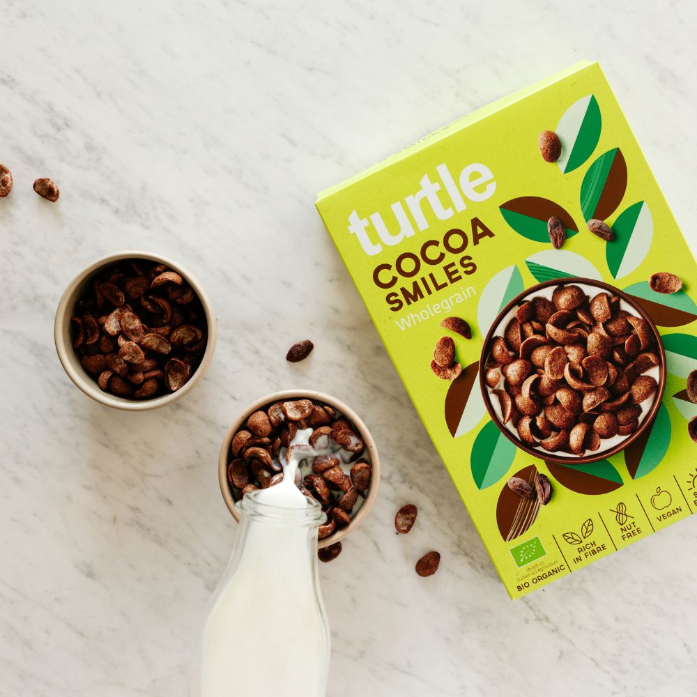 A bright yellow-green box of turtle Cocoa Smiles Wholegrain cereal is shown on a light marble surface, with bowls of chocolate cereal and a glass of milk nearby.
