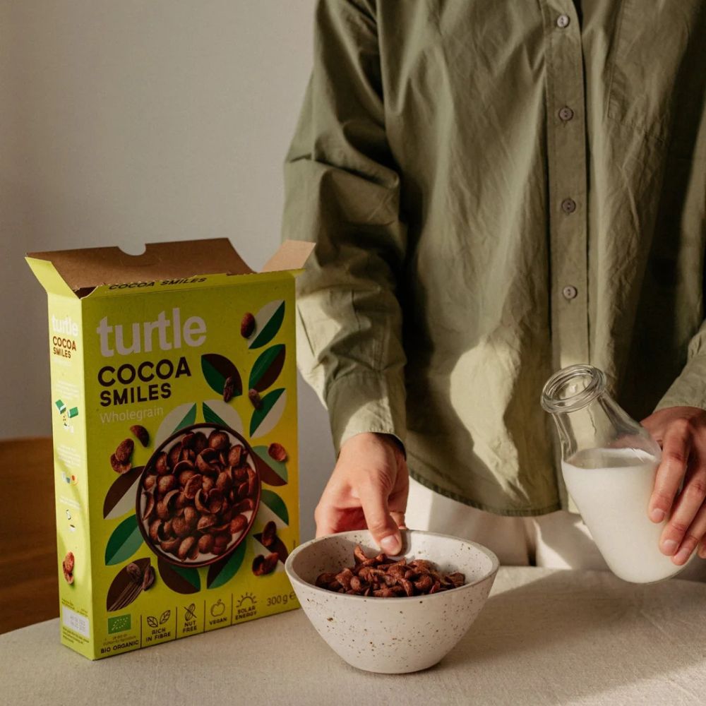 A person in an olive green shirt stands beside a box of Turtle Cocoa Smiles wholemeal cereal and a bowl of cereal, holding a glass of milk in a softly lit indoor setting.