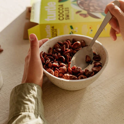 A person holds a speckled white bowl filled with chocolate-covered cereal pieces, using a metal spoon. A yellow box labeled Turtle Cocoa Snacks sits in the background on a beige table.