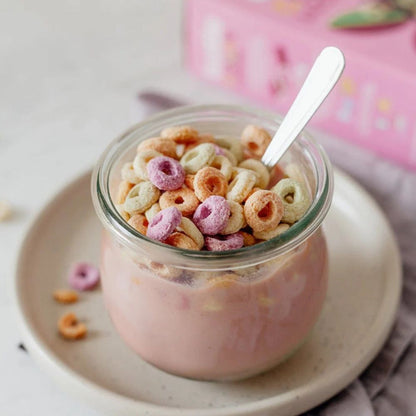 A clear glass jar filled with a pink yogurt or milk base and topped with colorful cereal loops, set on a beige plate with a spoon. A pink box is blurred in the background.