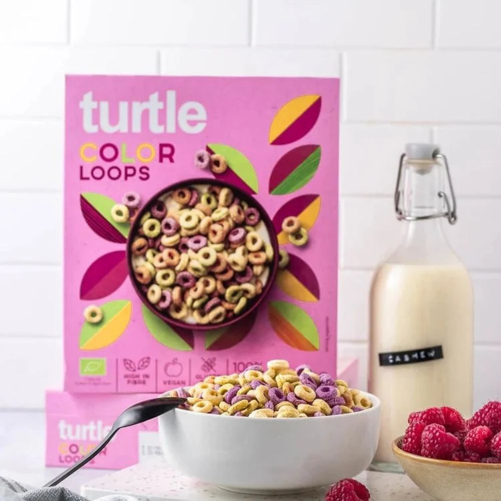 A bowl of Turtle Color Loops cereal in a white bowl with a spoon, set in front of its pink packaging box. A glass bottle of milk and a bowl of fresh raspberries are arranged on a white tiled countertop.
