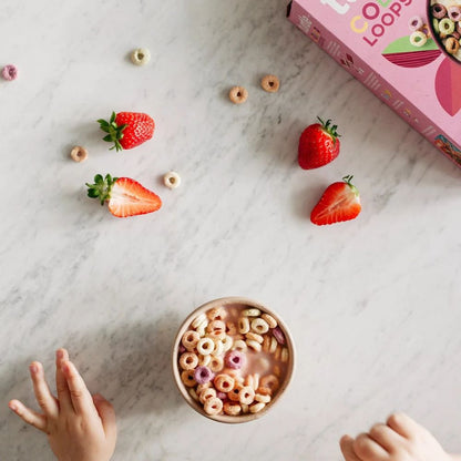 A styled flat-lay of a children's cereal box on a white marble surface, with a bowl of cereal and milk at the bottom, scattered strawberries, cereal pieces, and two small hands entering from the lower corners.
