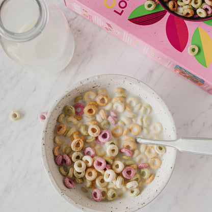 A top-down view of a white speckled bowl filled with milk and multicolored cereal loops, with a pink cereal box and a glass of milk on a white marble surface.