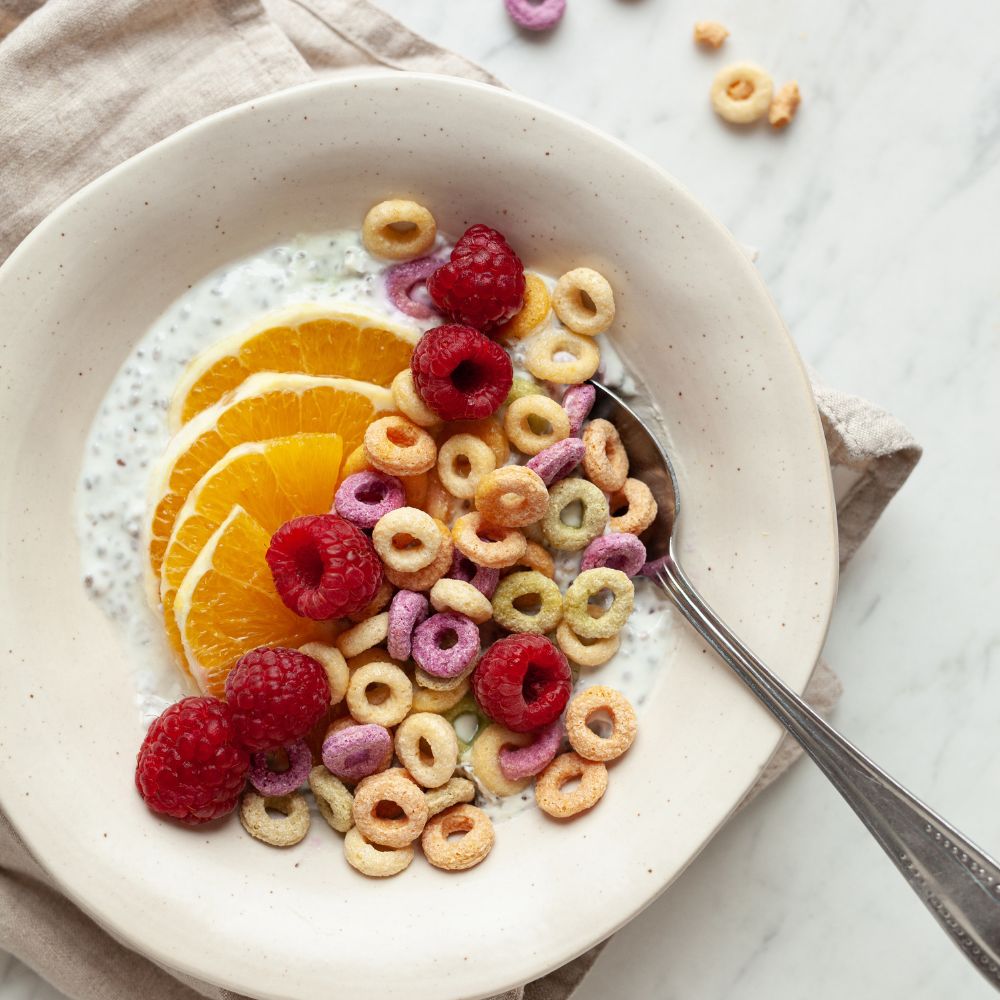 A top-down view of a breakfast cereal bowl on a marble surface with a beige cloth napkin. The bowl contains a creamy white drink, colorful ring-shaped cereal pieces in yellow, pink, and beige, sliced orange, and fresh red raspberries.