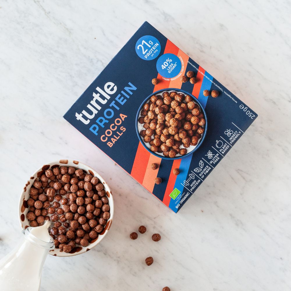 A box of Turtle Protein Cocoa Balls cereal is shown on a light marble surface next to a white bowl filled with the chocolate-coated balls and a milk pitcher.