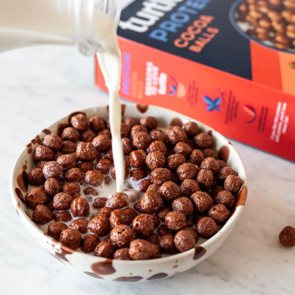 A bowl of chocolate protein cocoa balls cereal with milk being poured from a glass, with the red and black box of Tropicana Protein Cocoa Balls in the background on a light marble surface.