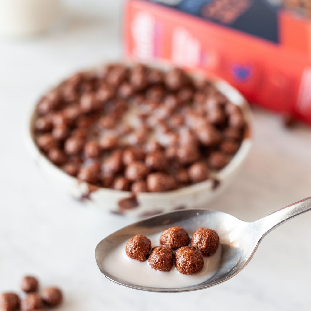 Chocolate cereal balls in milk, shown in a bowl with a spoonful in the foreground and the product packaging blurred in the background.
