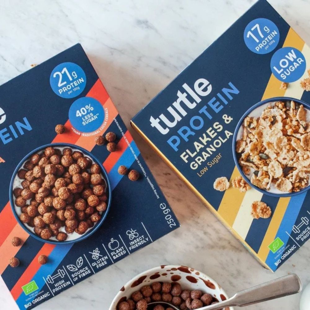 Two boxes of Turtle Protein cereal flakes and granola shown on a light marble surface, with a bowl of cereal and spoon in the foreground.