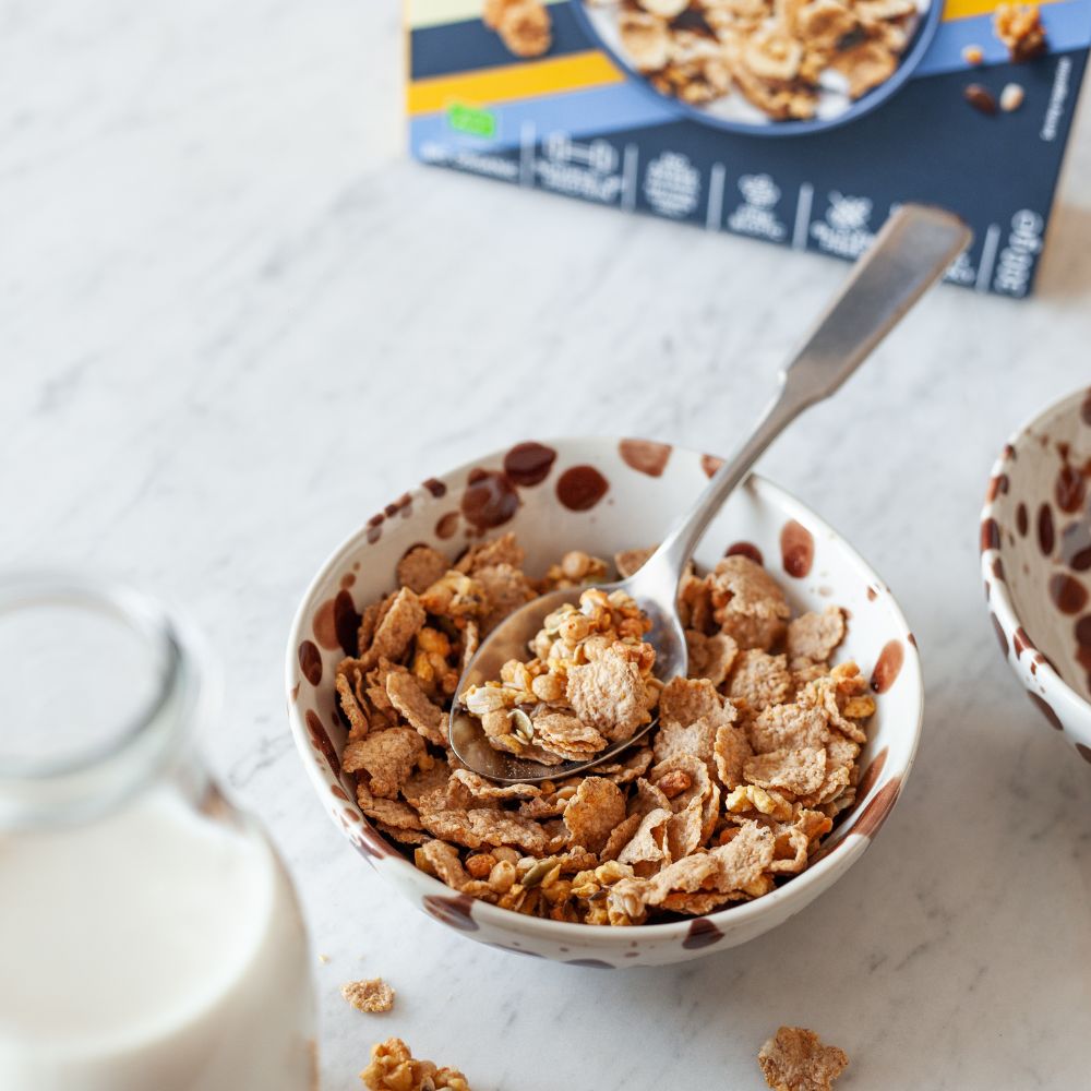 A bowl of granola cereal with a metal spoon, set on a light marble surface next to a glass of milk, with a cereal box blurred in the background.