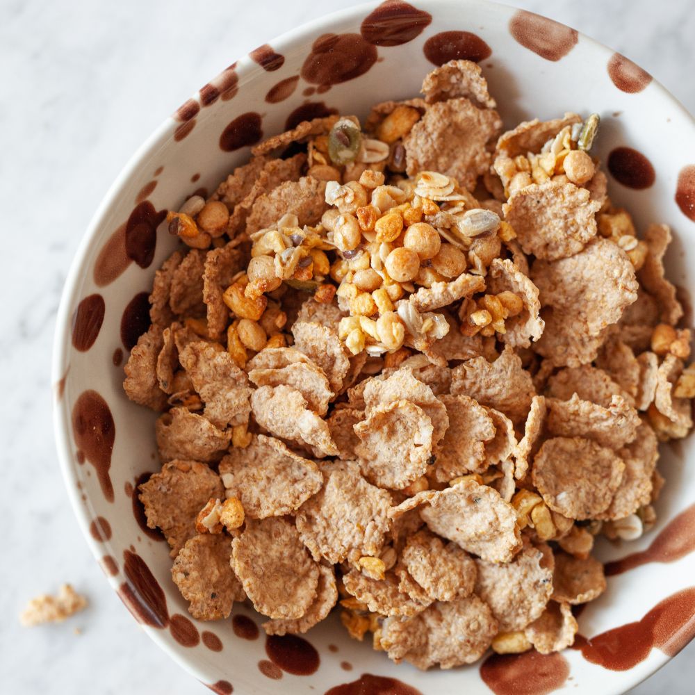 A top-down view of a bowl filled with a mixture of corn flakes, oats, and lentils, set against a light marble background.
