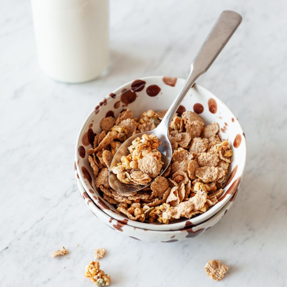 A bowl of cereal with a spoon on a light marble surface, with a blurred glass of milk in the background.