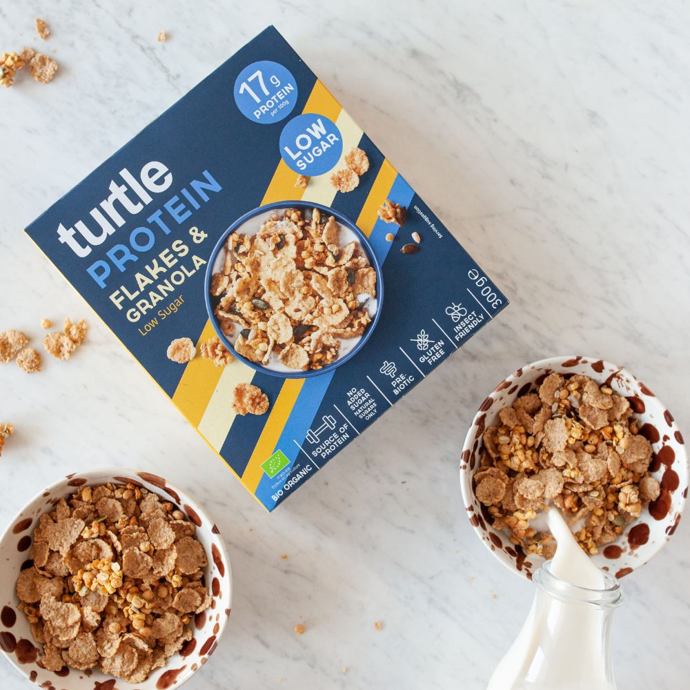 A box of Turtle Protein Flakes & Granola, Low Sugar, styled on a white marble surface with two bowls of the cereal and a glass of milk.