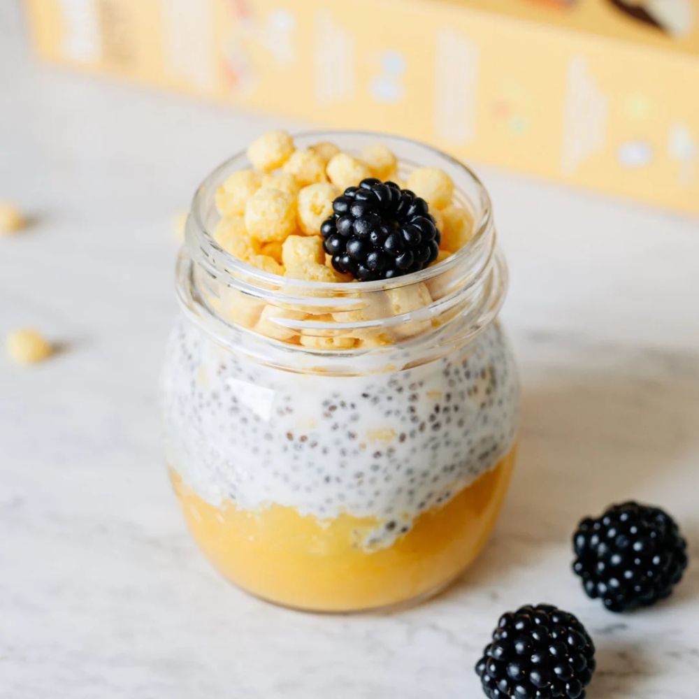 A clear glass mason jar filled with layered yellow-orange fruit pulp, white chia seed pudding, and topped with cereal pieces and a blackberry, set on a light marble surface with loose blackberries nearby.