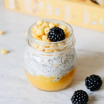 A clear glass mason jar filled with layered yellow-orange fruit pulp, white chia seed pudding, and topped with cereal pieces and a blackberry, set on a light marble surface with loose blackberries nearby.